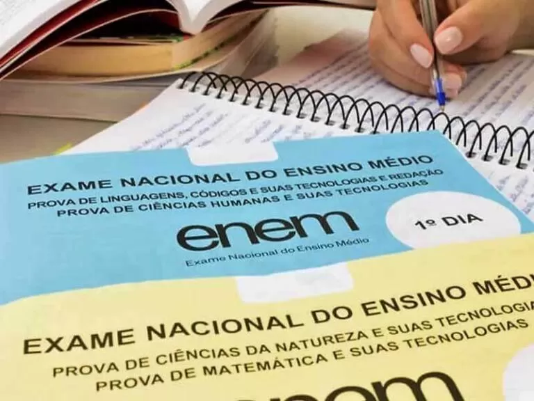 imagem de um caderno de prova do enem por cima de um caderno aramado e uma mao de uma moca escrevendo no caderno